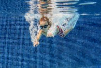 Junge im Grundschulalter in Schutzbrille schwimmt in blauem Pool unter Wasser mit Luftblasen — Stockfoto
