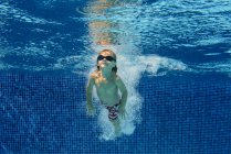 Elementary age boy in goggles swimming in blue pool underwater with air bubbles — Stock Photo