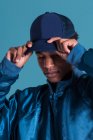 Trendiger ethnischer Mann mit glänzend blauer Bomberjacke und Mütze vor blauem Hintergrund — Stockfoto