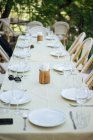 Belle table avec tissu léger et chaises en rotin autour ensemble avec des verres à vin vides et des assiettes à l'extérieur sur le trottoir avec des arbres et de l'herbe autour — Photo de stock