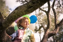 Хлопчик дошкільного віку сидить з закритими очима на дереві з повітряними кулями на сонячному світлі — стокове фото