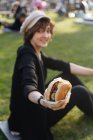 Улыбающаяся женщина показывает бургер, сидя на траве в парке — стоковое фото