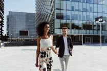Smiling elegant multiracial couple holding hands while walking in modern city — Stock Photo