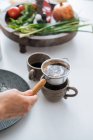 Coltivare le mani versando acqua calda dal bollitore in bella tazza di ceramica con piccolo filtro scoop in esso in piedi sulla tavola con ingredienti di cottura — Foto stock
