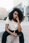 Вдумчивая афроамериканка, сидящая на городской улице и держащая смартфон — стоковое фото