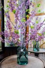 Hermosas ramitas largas recién cortadas con hojas verdes y flores púrpuras de pie en frasco de vidrio con agua en la mesa redonda de madera en la vieja habitación de mala calidad con espejos - foto de stock
