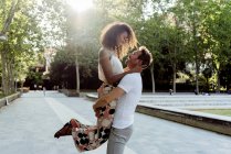 Homem levantando mulher rindo enquanto estava em pé no beco do parque no dia ensolarado — Fotografia de Stock