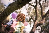 Low angle view of boy sitting on tree with balloons — Stock Photo