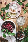Colocação plana de tigela de cerâmica cheia de cerejas e composta por folhas verdes, açúcar e limão em mesa rústica — Fotografia de Stock