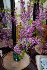Schöne frisch geschnittene lange Zweige mit grünen Blättern und lila Blüten stehen im Glas mit Wasser auf einem runden Holztisch im alten schäbigen Raum mit Spiegeln — Stockfoto