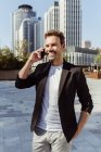 Улыбающийся элегантный мужчина улыбается разговаривая по телефону, стоя на улице современного города в солнечный день — стоковое фото
