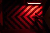 Vue latérale du mouvement figure floue d'un homme marchant dans un tunnel en direction opposée à de grandes flèches rouges et noires sur un mur éclairé par des lampes rouges — Photo de stock