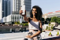 Elegant African-American woman posing for selfie while sitting on fence on city street on sunny day — Stock Photo
