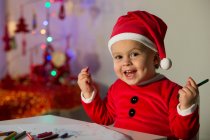 Happy little child in Christmas clothes drawing at table and looking at camera — Stock Photo