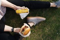 Mujer sentada en la hierba en el parque sosteniendo hamburguesa para llevar y pedazo de pastel - foto de stock