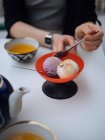 Vista de la cosecha de la persona sin rostro sentado en la mesa con té y cuchara de celebración con helado - foto de stock