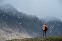 Вид сзади на самца в теплой одежде с рюкзаком в горах, стоящего на траве и смотрящего на зазубренный горный хребет, покрытый снегом, и вершины, скрытые в облаках — стоковое фото