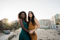 Trendige junge, vielfältige Frauen, die sich im Sonnenlicht umarmen, mit Stadtbild im Hintergrund — Stockfoto