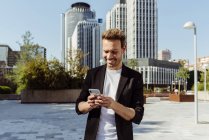 Элегантный улыбающийся парень просматривает смартфон на улице современного города — стоковое фото