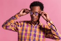 Trendy black man wearing colorful checkered shirt with shiny pink sunglasses standing on pink background — Stock Photo
