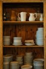 Beautiful rustic wooden plank dish cabinet with porcelain pitchers and ceramic plates and saucers standing in piles on shelves — Stock Photo