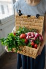 Mulher de colheita em camisa e saia segurando em mãos cesta com tampa aberta cheia de tomates frescos brilhantes, pimenta, rabanete e potherbs — Fotografia de Stock