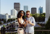 Laughing multiracial couple using smartphone while standing on background of modern city — Stock Photo