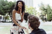 Caucasian man putting engagement ring on finger of black woman while proposing near fountain in park — Stock Photo