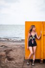 Slender woman in shorts and top leaning on orange metal wall while standing on sandy beach at seaside — Stock Photo