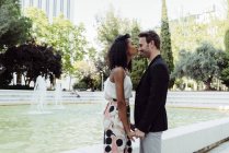 Charmant couple multiracial embrassant tout en se tenant près de la fontaine dans le parc — Photo de stock