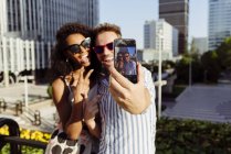 Cheerful multiracial couple posing for selfie while standing on background of modern city — Stock Photo