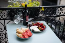 Бутылка вина и хрупкие бокалы с высокими стеблями, стоящими на столе с нарезанными помидорами, хлебом, сыром и ягодами, лежащими на тарелках с декоративным забором и растениями на заднем плане — стоковое фото