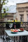 Eine Flasche Wein und zarte Gläser mit hohen Stielen stehen auf dem Tisch mit geschnittenen Tomaten, Brot, Käse und Beeren liegen auf Tellern mit Zierzäunen und Pflanzen im Hintergrund. — Stockfoto