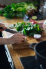 Crop female hands chopping green leaves of herbs on wooden cutting board on table with stove nearby — Stock Photo