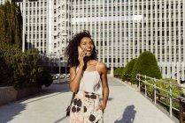Elegante mujer afroamericana hablando en un teléfono inteligente mientras camina por la calle de la ciudad en un día soleado - foto de stock
