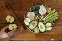 Жіночі руки нарізають яблука і готують здорову тарілку з зеленими фруктами та овочами на дерев'яній поверхні — стокове фото