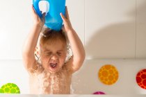 Cute little happy kid sitting in tub with colorful round decoration and splashing water on head with closed eyes — Stock Photo
