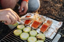 Human hands with tongs checking salmon and zucchini pieces on foil on grill grid — Stock Photo
