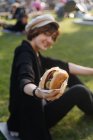 Улыбающаяся женщина показывает бургер, сидя на траве в парке — стоковое фото