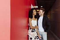 Beau homme appuyé sur le mur rouge et flirtant avec la femme afro-américaine dans le couloir de construction — Photo de stock