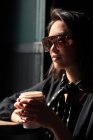 Femme élégante dans les lunettes de soleil et le foulard tenant tasse de papier de café et penché sur la table — Photo de stock