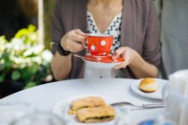 Female hands holding red ceramic polka-dotted mug of tea on saucer on garden table — Stock Photo