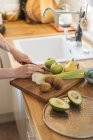 Manos femeninas rebanando ingredientes y preparando un plato saludable con frutas y verduras verdes en la superficie de madera - foto de stock