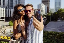 Cheerful multiracial couple posing for selfie while standing on background of modern city — Stock Photo