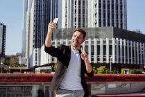 Веселый мужчина в повседневной одежде показывает победный жест и позирует для селфи в современном городе — стоковое фото