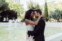Elegant multiracial couple embracing near fountain in city park — Stock Photo