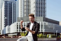 Excited man holding smartphone and rejoicing over victory while standing on street of modern city — Stock Photo