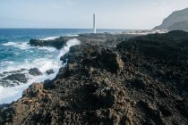 Мальовничий вид на сірі скелясті скелі з вежею маяка на фоні берегової лінії океану з сильними хвилями — стокове фото