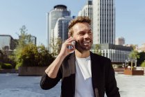 Sonriendo elegante hombre sonriendo hablando por teléfono mientras está parado en la calle de la ciudad moderna en el día soleado - foto de stock