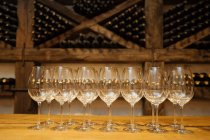 Fila di eleganti bicchieri lucidi in piedi su tavolo di legno in cantina con bottiglie di vino su scaffali sullo sfondo — Foto stock